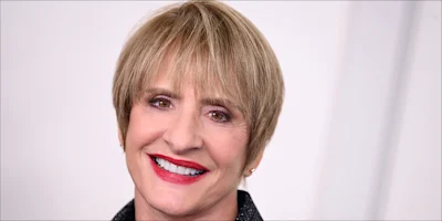 A person with short light brown hair, wearing a dark jacket, smiles in front of a plain light background.