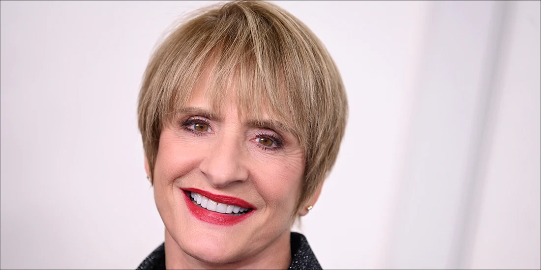 A person with short light brown hair, wearing a dark jacket, smiles in front of a plain light background.
