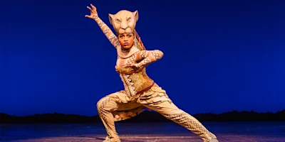 Performer in a lioness costume strikes a dynamic pose on stage against a plain blue background.