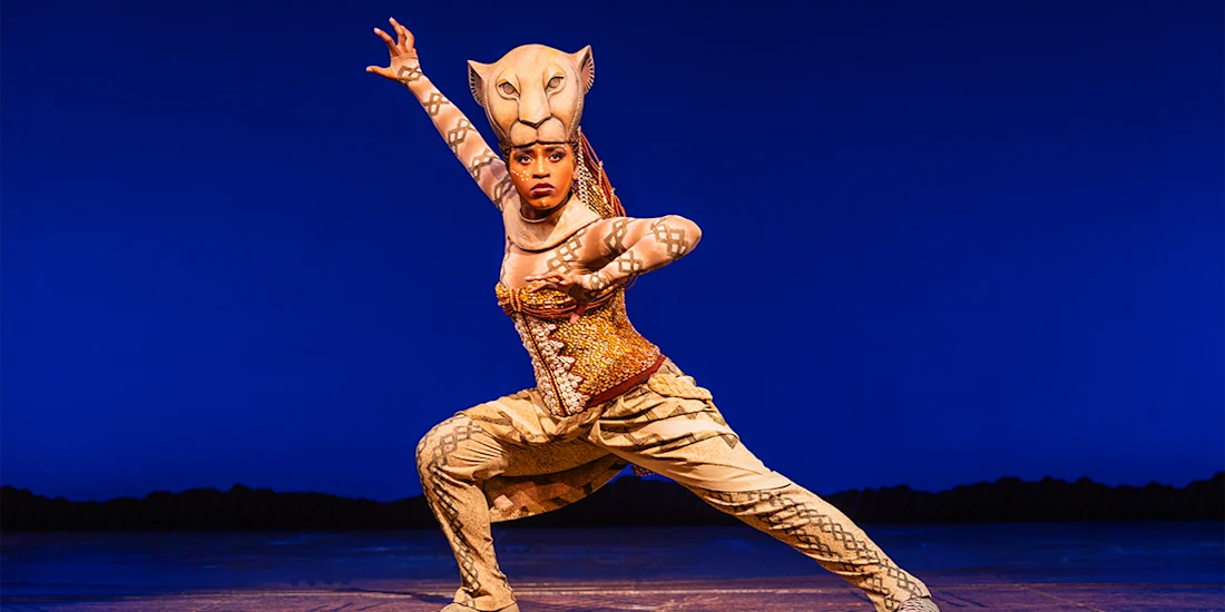 Performer in a lioness costume strikes a dynamic pose on stage against a plain blue background.