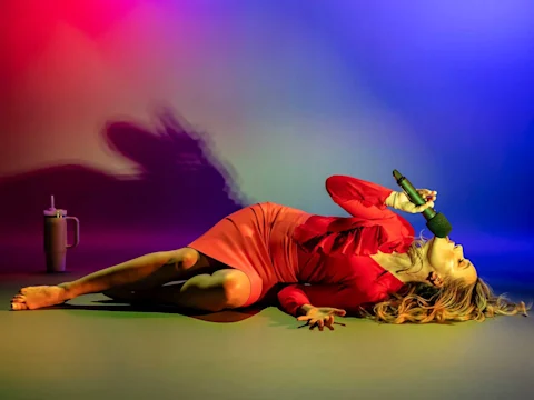 A woman in a red dress lies on the floor singing into a microphone, with a large water bottle nearby, under colorful stage lighting.
