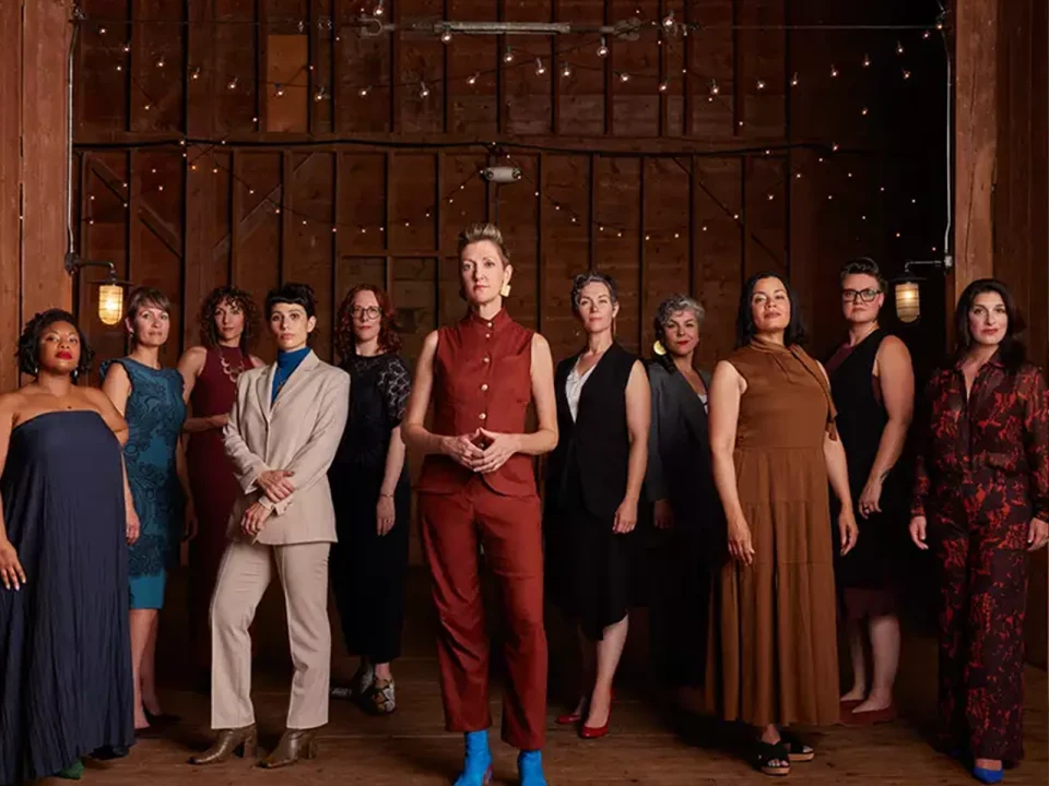 A diverse group of twelve people stands in a wooden room with string lights, wearing formal and casual attire.