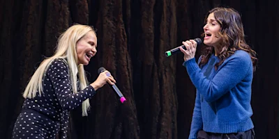 Two women stand facing each other, holding microphones and singing passionately on stage in front of a dark, textured backdrop.