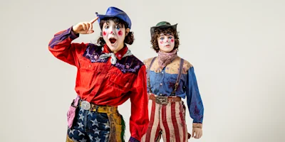 Two people dressed as clowns in colorful Western-style outfits stand against a plain background, one in a red shirt and blue hat, the other in a blue shirt and green hat.