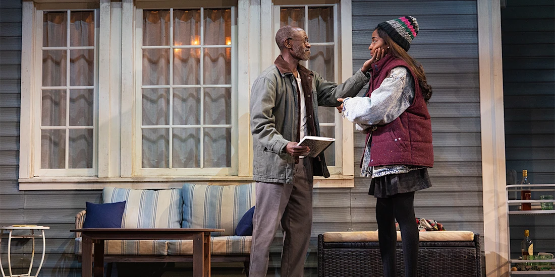 A man and a woman stand on a porch set; the man touches the woman’s face while holding a notebook, and the woman, bundled in winter clothing, looks at him.
