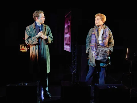 Two people stand on a dimly lit stage. One wears a trench coat and glasses; the other has short hair and a crossbody bag. The background is dark with hints of red light.