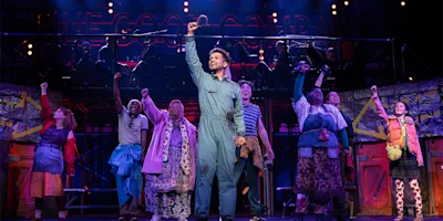 A group of performers on stage, dressed in colorful costumes, stand with fists raised, with one person holding a microphone at the center under bright stage lights.