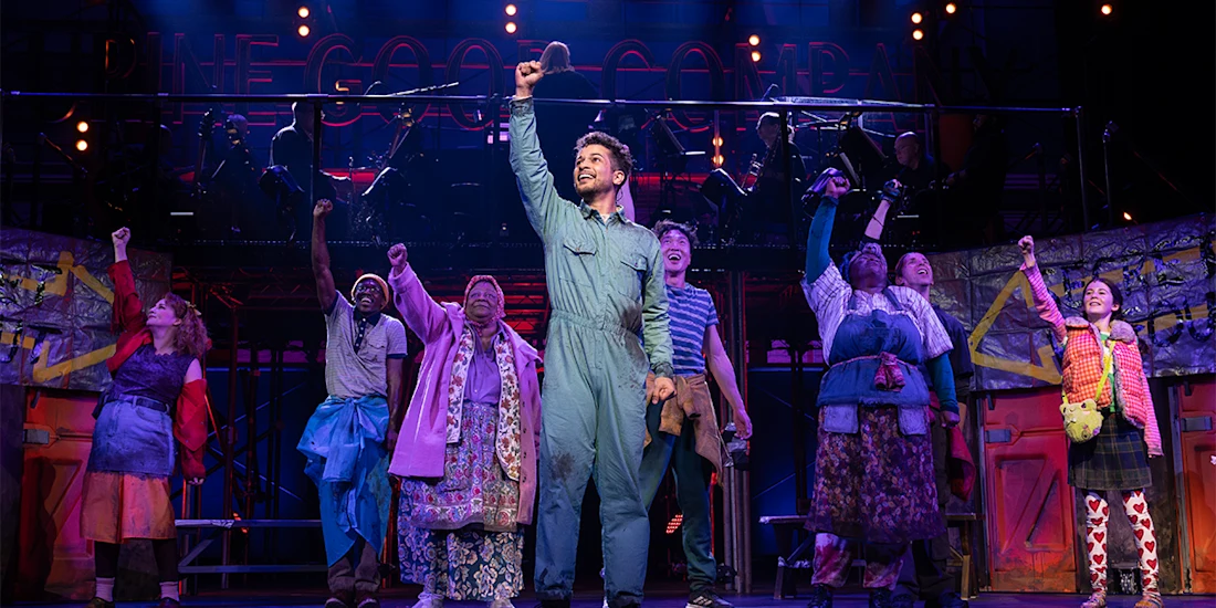 A group of performers on stage, dressed in colorful costumes, stand with fists raised, with one person holding a microphone at the center under bright stage lights.