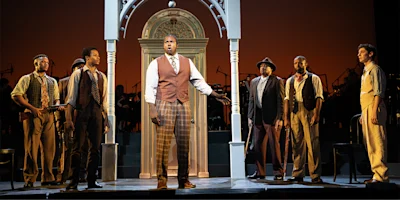Seven men in period costumes stand on stage in front of a large decorative archway, performing in a theatrical production with musicians visible in the background.