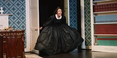A woman in a long black dress with a white collar energetically enters a room with patterned walls and bookshelves during a stage play.