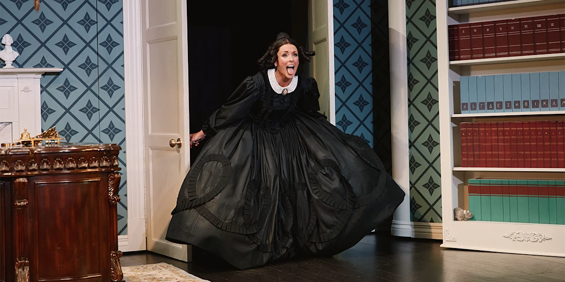 A woman in a long black dress with a white collar energetically enters a room with patterned walls and bookshelves during a stage play.