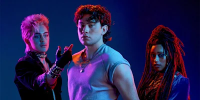 Three people pose against a dark background with colored lighting. The person on the left has spiked pink hair, the center person wears a gray sleeveless shirt, and the right person has long braided hair.