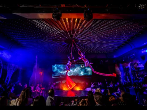 Production photo of MOONRISE: An Immersive Theatrical Experience at The Paradise Club in New York, showing two aerial performers suspended in mid-air by red ribbons perform above a stage with an audience seated around them.