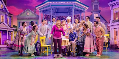 A woman stands center stage holding a pie, surrounded by smiling actors in colorful costumes, with a gazebo and houses in the background.