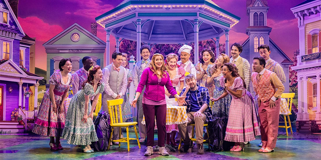 A woman stands center stage holding a pie, surrounded by smiling actors in colorful costumes, with a gazebo and houses in the background.