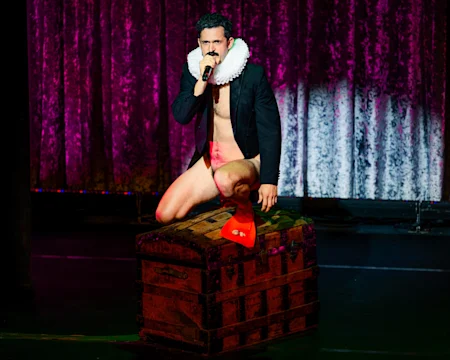 A man wearing a ruff collar, blazer, and swim fins kneels on a large wooden chest, holding a microphone, with a purple velvet curtain in the background.