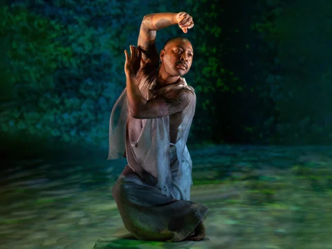 A person with tattoos performs a dance on one knee, wearing a light-colored sleeveless outfit against a green, textured background.