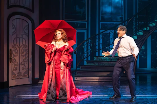 Two actors on stage: one in a red gown holding a red umbrella, the other in a white shirt, tie, and glasses, appearing surprised, with a staircase in the background.