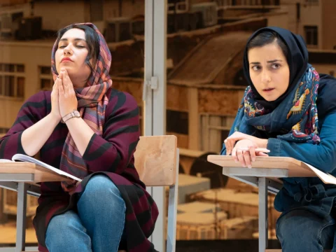 Two women wearing headscarves sit at school desks; one is praying while the other looks worried. They are in an indoor setting with a blurred background.