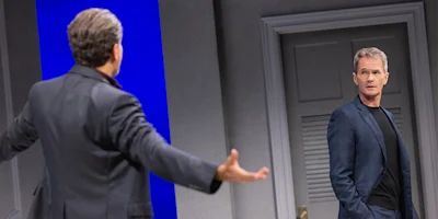 Two men in suits stand in a modern room; one faces away with arms outstretched, while the other looks at him, appearing surprised or concerned.