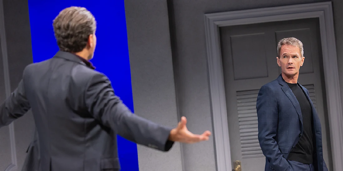 Two men in suits stand in a modern room; one faces away with arms outstretched, while the other looks at him, appearing surprised or concerned.
