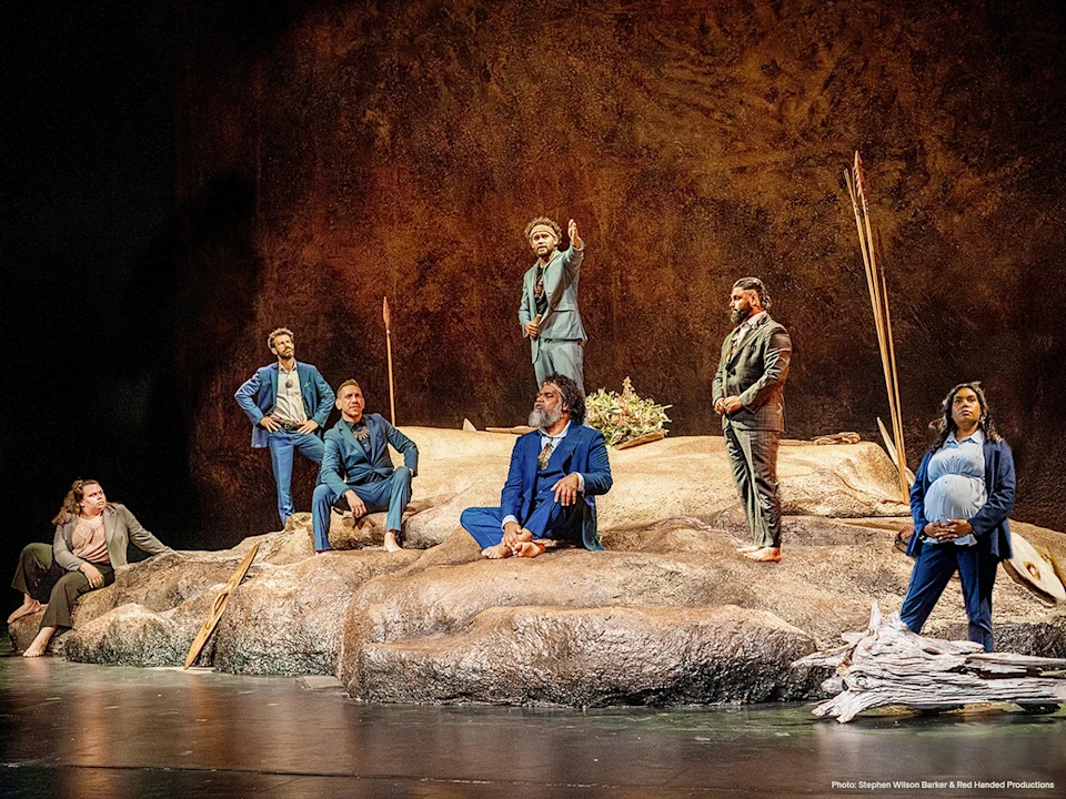 Seven actors in suits stand and sit on a rocky stage set, some holding sticks, suggesting a dramatic theater performance.
