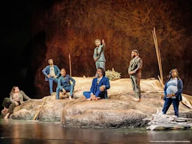 Seven actors in suits stand and sit on a rocky stage set, some holding sticks, suggesting a dramatic theater performance.