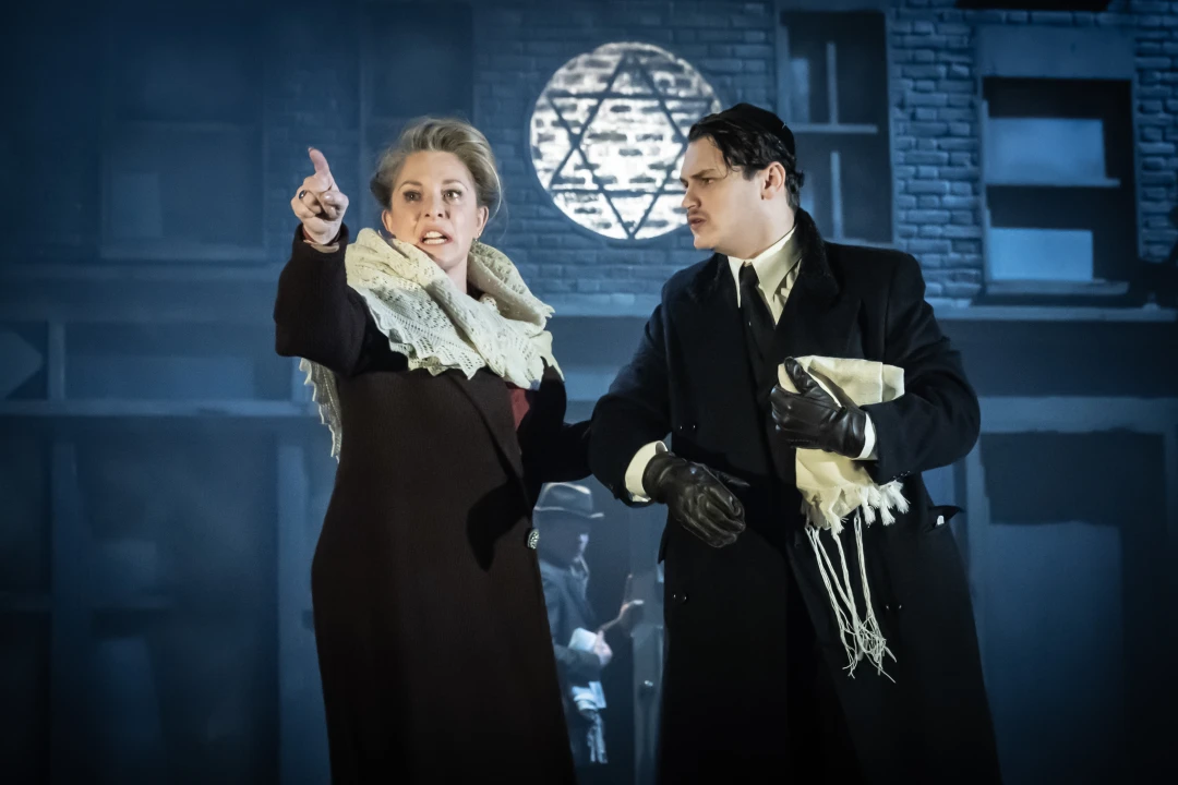 Production image of The Merchant of Venice in London, featuring Tracy Ann Oberman as Shylock with supporting ensemble member.