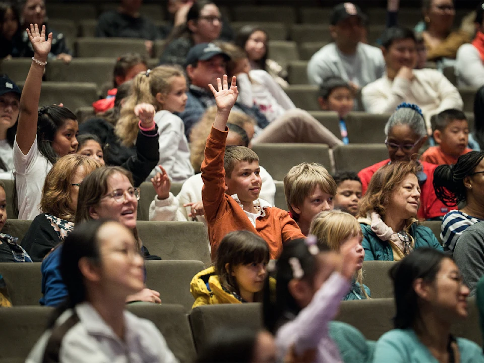 A diverse group of children and adults sit in an auditorium; several children in the center raise their hands, engaging with the speaker.