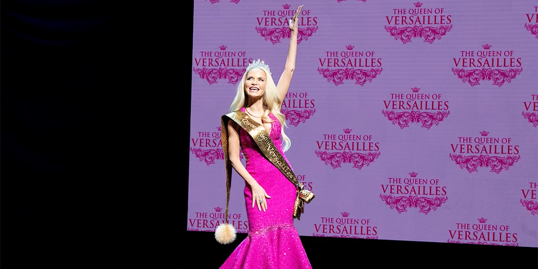 A woman in a pink gown, sash, and crown poses with one arm raised in front of a screen displaying "The Queen of Versailles" logo.