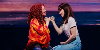 Two women sit facing each other, making a pinky promise, with warm lighting highlighting their expressions and colorful clothing.