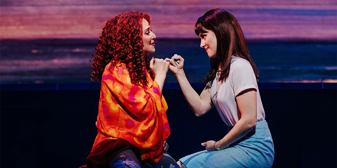 Two women sit facing each other, making a pinky promise, with warm lighting highlighting their expressions and colorful clothing.