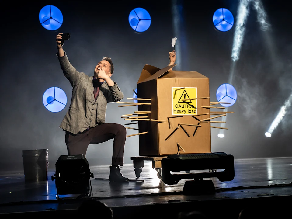 A person kneels taking a selfie on stage next to a large box labeled "Caution Heavy Load" with arms holding cards sticking out. Industrial lights are visible in the background.