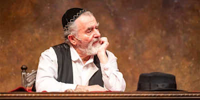 An older man with a gray beard, wearing a kippah and vest, sits at a table resting his chin on his hand, looking thoughtful. A black hat is on the table beside him.