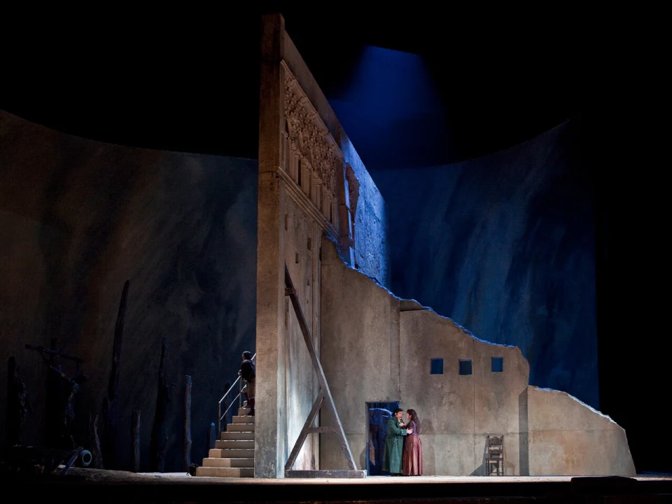 Production Photo of Il Trovatore in New York, showing two people in period costumes stand near the corner of a tall, weathered set piece on stage. The scene is dimly lit with a blue backdrop and a staircase leading up the structure.