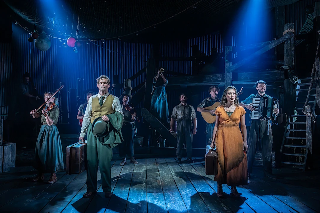 Cast members in period costumes perform on a wooden stage, with some carrying suitcases and instruments under blue spotlights.