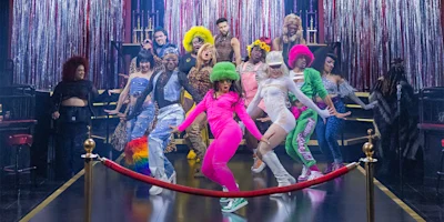 A group of performers in colorful, flamboyant costumes dance energetically on a stage with red curtains and sparkly silver tinsel in the background.