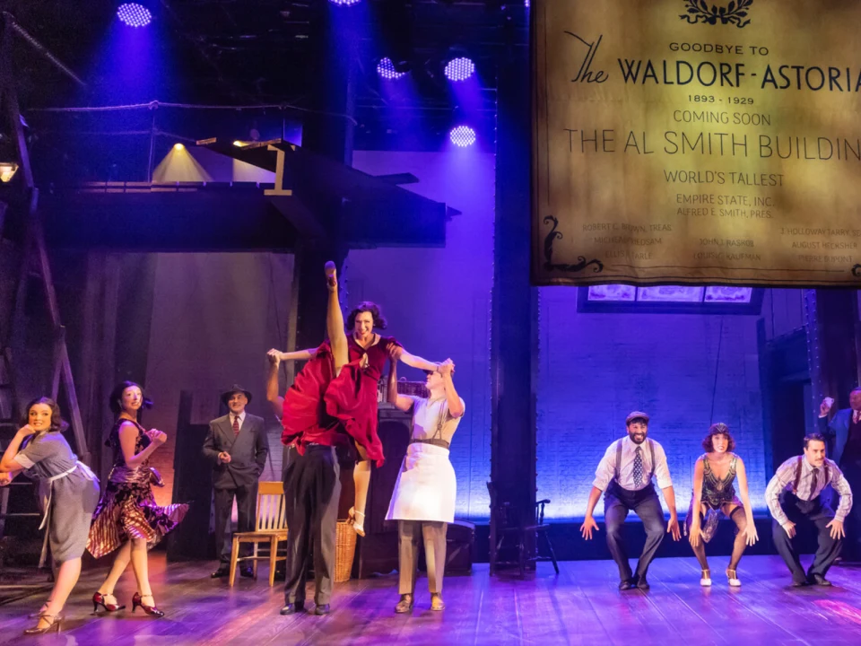 Actors perform a lively dance routine on stage, with a woman in a red dress being lifted in the air. A backdrop ad for the Waldorf-Astoria and Al Smith Building is visible.