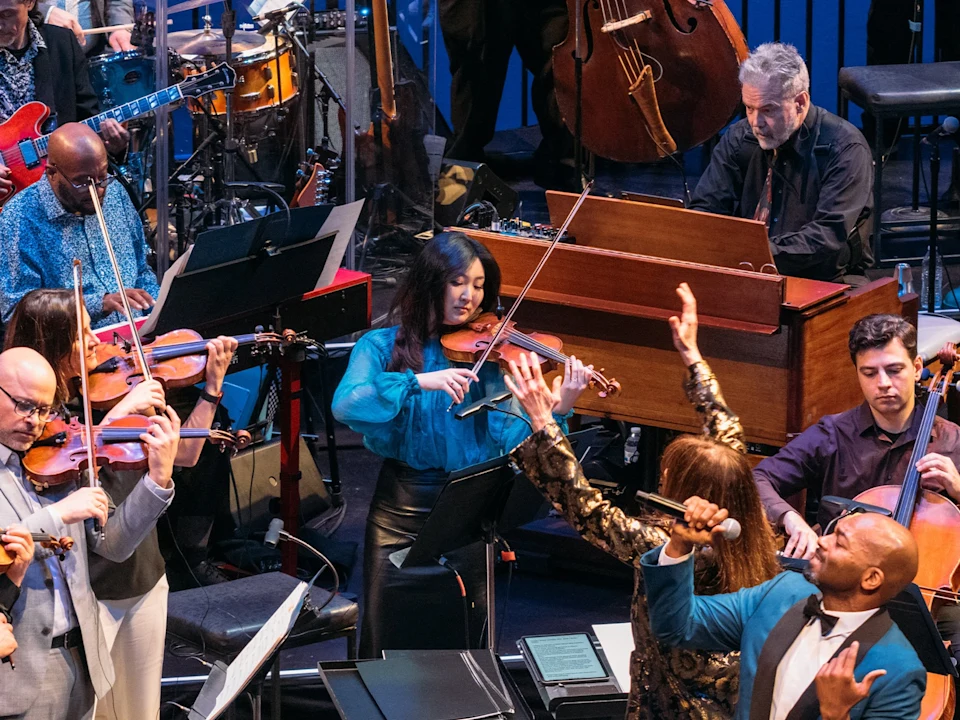 A live orchestra with string musicians, a woman playing violin, a man at a keyboard instrument, and two singers performing on stage.