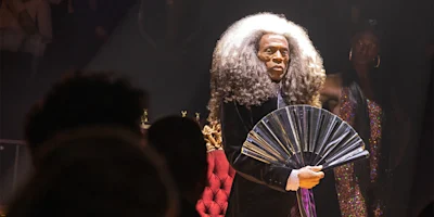 Person with long, voluminous gray hair holds a shiny folding fan while standing under a spotlight, with people and a red chair in the background.