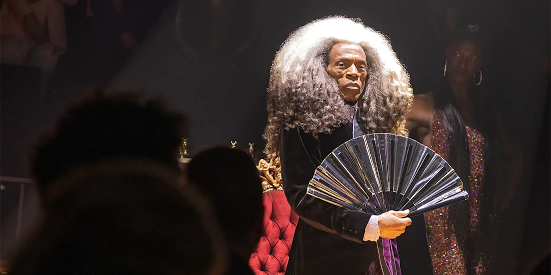 Person with long, voluminous gray hair holds a shiny folding fan while standing under a spotlight, with people and a red chair in the background.