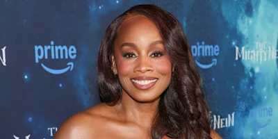 A woman with long, wavy hair smiles at the camera on a blue-themed event backdrop featuring Amazon Prime and The Wheel of Time logos.