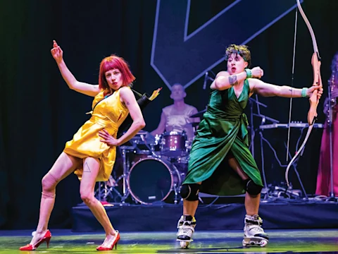 Two performers in colorful dresses pose dramatically on stage, one in yellow with red hair and heels, the other in green with a bow and rollerskates; a drummer plays in the background.