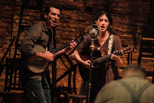 A man playing a banjo and a woman playing an acoustic guitar and singing into a vintage microphone on a stage.