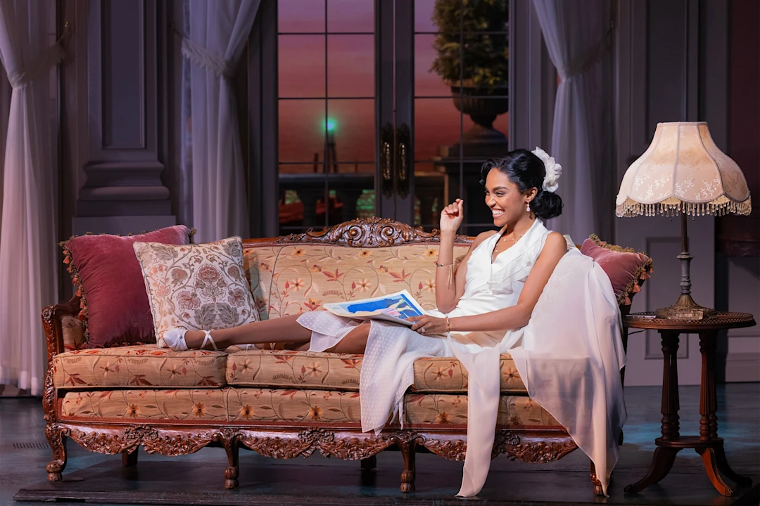 A woman in a white dress sits on an ornate sofa, smiling and holding a magazine. The scene is indoors with elegant decor and a large window showing a sunset view.