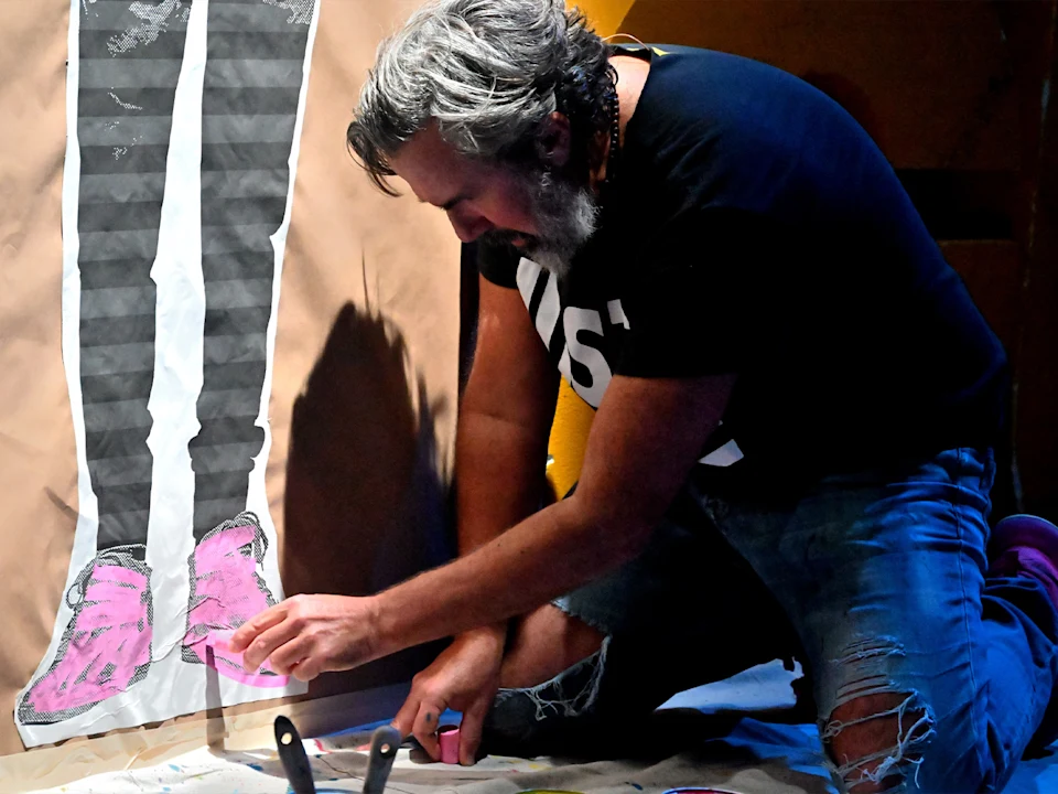 A man kneels on the floor, drawing or painting pink shoes on a large artwork depicting a pair of legs on brown paper.