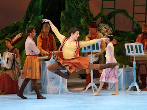 Production photo of American Ballet Theatre: The Winter’s Tale in washington, showing a male dancer leaps mid-air in a theatrical performance, surrounded by musicians and other performers in colorful costumes on a stage with a nature-themed backdrop.
