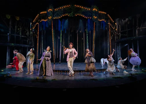 Actors in colorful costumes perform on a stage set resembling a forest, with a lit arch structure and trees in the background.