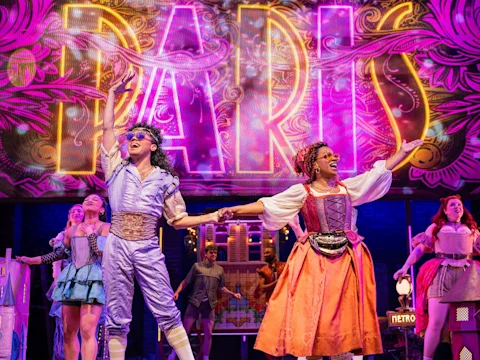 Performers in colorful costumes sing and hold hands on stage in front of a bright "PARIS" sign during a theatrical musical scene.