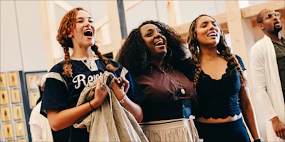 Three women singing with their mouths open, standing in a rehearsal space with a man in the background.